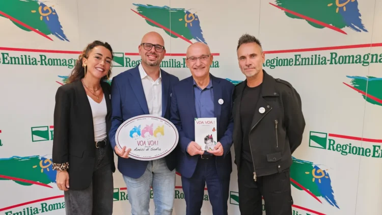 Giulia Ferrari, Guido De Barros, Massimo Fabi e Nek posano insieme davanti al fondale della Regione Emilia-Romagna; Guido tiene il cartello di Voa Voa e Fabi un libro.