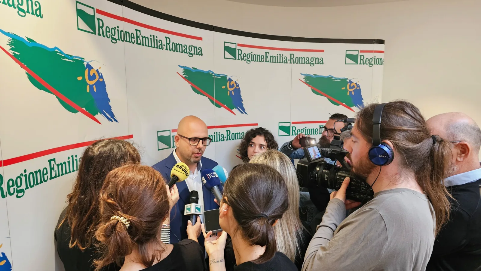 Guido De Barros, presidente di Voa Voa, risponde ai giornalisti circondato da microfoni e telecamere nella sede della Regione Emilia-Romagna.